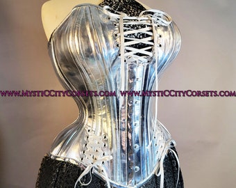 May include: A silver metallic corset with white lacing in the front. The corset has a boned structure and a smooth, shiny finish. The website address "www.mysticcitycorsets.com" is printed on the bottom of the corset.