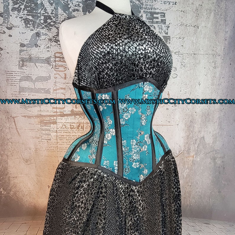 May include: A turquoise and black corset with a floral pattern and a silver halter top. The corset has a boned structure and a lace-up back. The website address "www.mysticcitycorsets.com" is printed on the front of the corset.