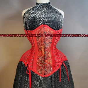 May include: A red corset with a gold and red peacock feather design. The corset has a mesh panel on the back and is laced up the front. The corset is shown on a mannequin with a black and silver snake skin print halter top.