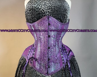Può includere: Un corsetto in broccato viola e argento con un pannello in rete nera e lacci in nastro viola. Il corsetto presenta un motivo floreale decorativo ed è stretto in vita. L'indirizzo del sito web "www.mysticcitycorsets.com" è stampato sul davanti del corsetto.