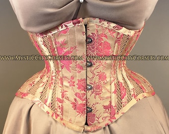 Può includere: Un corsetto floreale dorato e rosa con un pannello in rete sul davanti. Il corsetto ha una chiusura a busc ed è realizzato in tessuto satinato. Il corsetto è mostrato su una modella che indossa un abito beige.