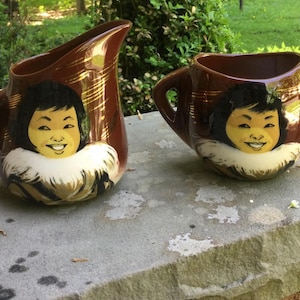 May include: A brown ceramic pitcher and sugar bowl set. Each piece features a smiling face with black hair and a white fur collar. Gold accents adorn the pitcher and bowl, adding a touch of elegance to the set. The set is placed on a stone surface.