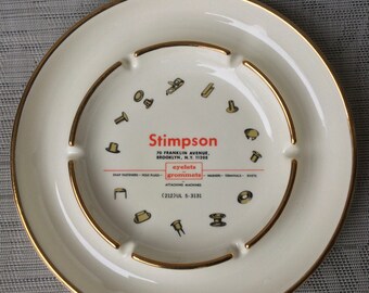 May include: A round white ceramic ashtray with a gold-trimmed rim and inner ring. The center features the text Stimpson, 70 Franklin Avenue, Brooklyn, N.Y. 11205, eyelets grommets, and (212)UL 5-3131, surrounded by small illustrations of metal fasteners.