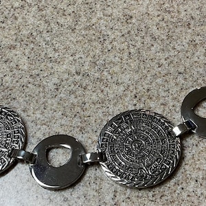 May include: A silver-toned metal bracelet featuring three circular medallions with intricate embossed calendar-style designs, alternating with three plain polished silver-toned circular links, connected by small metal hinges.