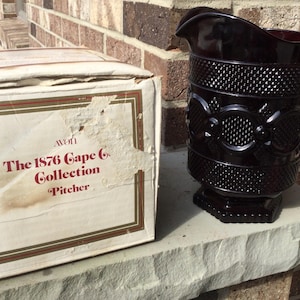 May include: A vintage, dark red glass pitcher from the Avon 1876 Cape Cod Collection. The pitcher has a textured pattern and a handle. The box next to it is labeled "The 1876 Cape Cod Collection Pitcher."