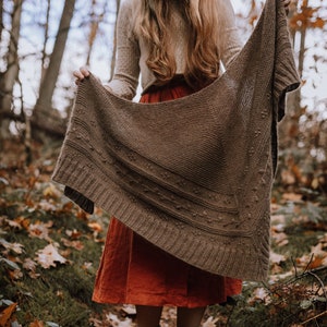 May include: A brown knitted shawl with textured patterns and a triangular shape. The shawl is held up in a forest setting, with a red dress and boots visible. The shawl appears to be handmade.
