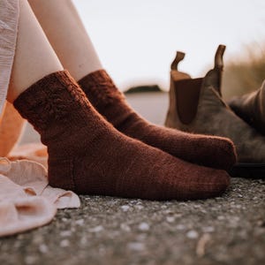 May include: A pair of brown knitted socks with a cable knit pattern. The socks are worn on a person's feet and are resting on a gravel surface.