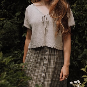 Könnte beinhalten: Eine Frau trägt ein hellbeiges, gestricktes Crop-Top mit einem Bindedetail am Ausschnitt und einen braunen und grünen karierten Rock.