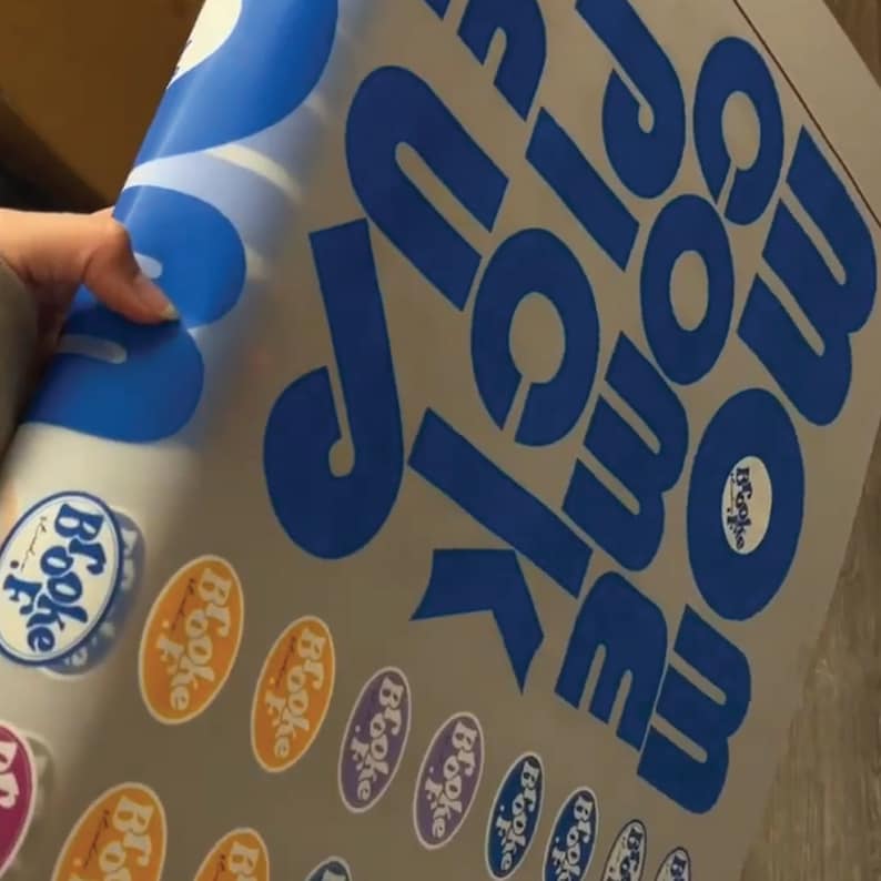 A hand holding a large sticker sheet featuring bold blue text reading 'Mom Come Pick Me Up' and multiple small oval stickers with 'Brooke' in various colors. The sheet appears glossy and ready for transfer