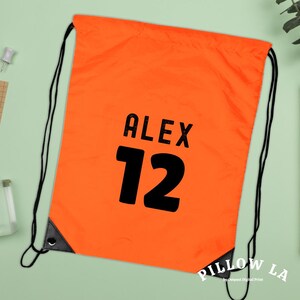 K&ouml;nnte beinhalten: Leuchtend orangefarbener Kordelzugbeutel mit schwarzen Kordeln und verst&auml;rkten Ecken. Der Beutel zeigt den Namen "ALEX" &uuml;ber der Zahl "12" in fetter schwarzer Schrift. Die untere rechte Ecke tr&auml;gt den wei&szlig;en Schriftzug "PILLOW LA".