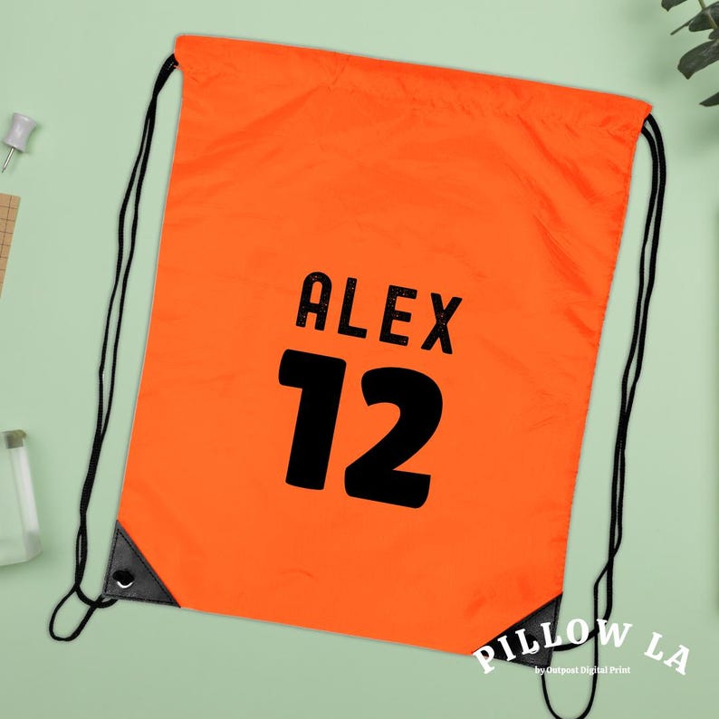 K&ouml;nnte beinhalten: Leuchtend orangefarbener Kordelzugbeutel mit schwarzen Kordeln und verst&auml;rkten Ecken. Der Beutel zeigt den Namen "ALEX" &uuml;ber der Zahl "12" in fetter schwarzer Schrift. Die untere rechte Ecke tr&auml;gt den wei&szlig;en Schriftzug "PILLOW LA".