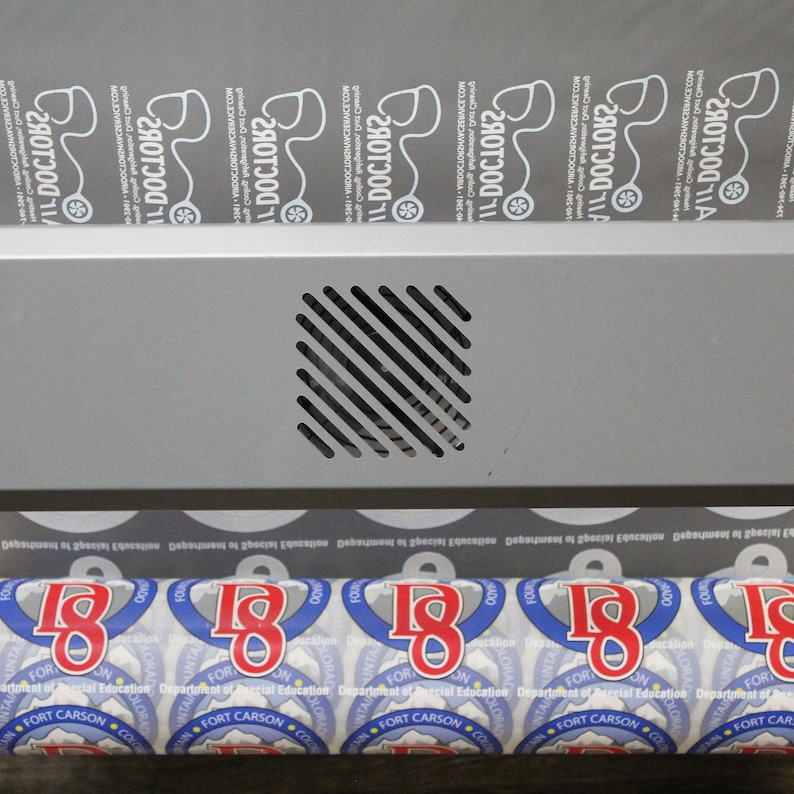 A roll of DTF prints in production, showing multiple logos for different businesses, including '11 Doctors' and 'Fort Carson Department of Special Education.' The sheet is being processed through a printer.