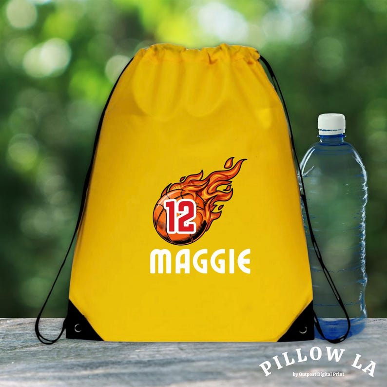 K&ouml;nnte beinhalten: Ein gelber Kordelzugbeutel mit einem Basketball-Design in Flammen, der Nummer 12 und dem Namen "Maggie" in Wei&szlig;. Rechts steht eine transparente Wasserflasche mit blauem Deckel. Der Beutel hat schwarze Kordeln.