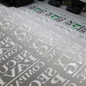 A sheet of glossy white vinyl lettering being printed, featuring reversed text in a bold serif font for a security company. The print has a clean, professional finish.