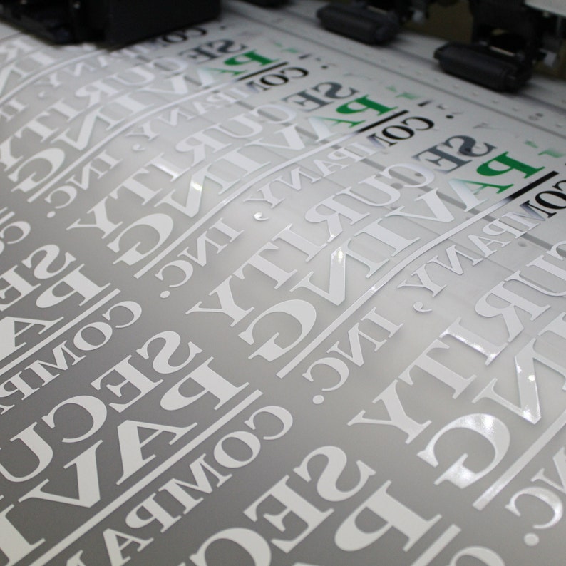 A sheet of glossy white vinyl lettering being printed, featuring reversed text in a bold serif font for a security company. The print has a clean, professional finish.