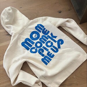 A white hoodie with a large blue text design reading 'Mom Come Pick Me Up' and a small circular logo on the back. The hoodie is laid flat on a wooden floor, showcasing the printed design