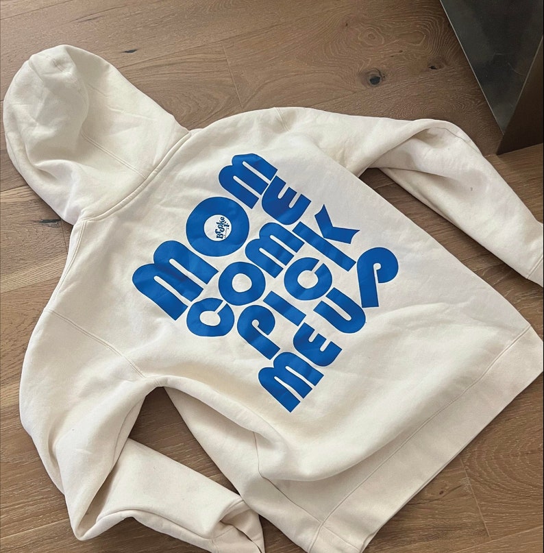 A white hoodie with a large blue text design reading 'Mom Come Pick Me Up' and a small circular logo on the back. The hoodie is laid flat on a wooden floor, showcasing the printed design