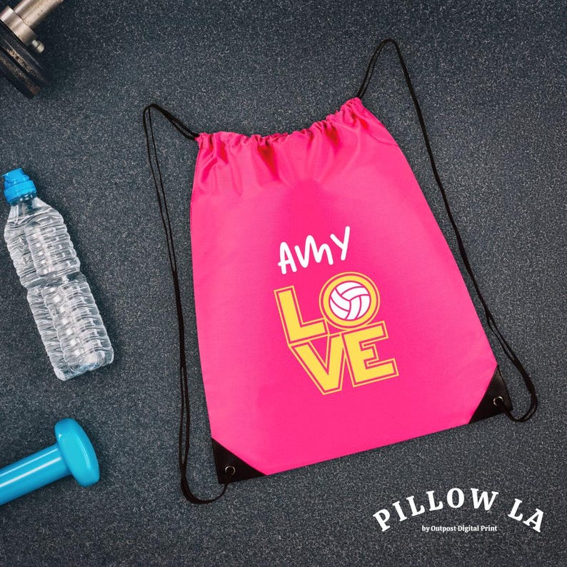 K&ouml;nnte beinhalten: Ein pinkfarbener Kordelzugbeutel mit den Worten "AMY LOVE" und einer Volleyballgrafik in Gelb und Wei&szlig;. Eine Wasserflasche und eine blaue Hantel liegen neben der Tasche. Die Tasche hat schwarze Kordelz&uuml;ge.