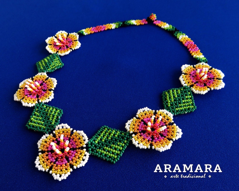 May include: A beaded necklace with a floral design. The necklace is made with white, yellow, orange, pink, and green beads. The necklace is on a blue background. The text "ARAMARA arte tradicional" is at the bottom of the image.