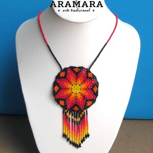 May include: A red, orange, and yellow beaded necklace with a flower design and black beads. The necklace is on a white mannequin bust. The text "ARAMARA arte tradicional" is above the necklace.