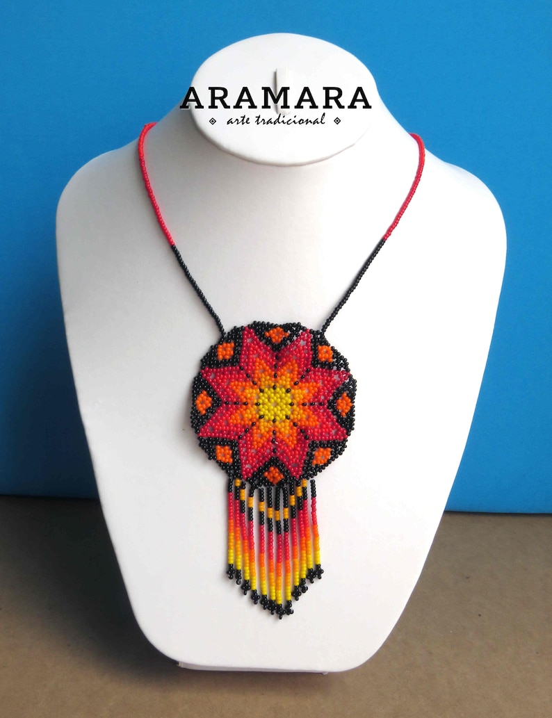 May include: A red, orange, and yellow beaded necklace with a flower design and black beads. The necklace is on a white mannequin bust. The text "ARAMARA arte tradicional" is above the necklace.