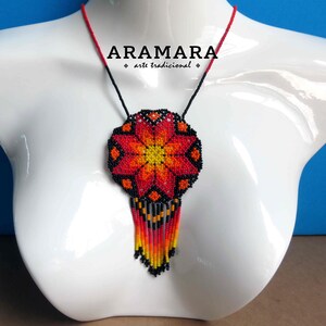 May include: A beaded necklace with a large, red, orange, and yellow flower pendant. The pendant has a black background and is surrounded by a black beaded fringe. The necklace is on a white mannequin bust. The text "ARAMARA - arte tradicional" is visible on the necklace.