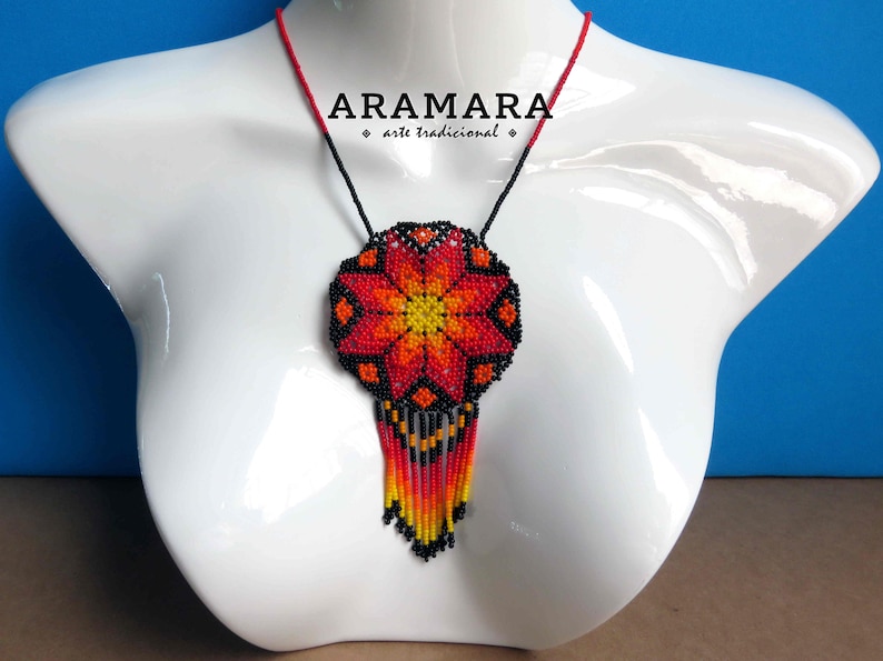 May include: A beaded necklace with a large, red, orange, and yellow flower pendant. The pendant has a black background and is surrounded by a black beaded fringe. The necklace is on a white mannequin bust. The text "ARAMARA - arte tradicional" is visible on the necklace.