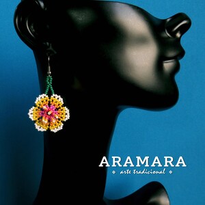 May include: A pair of beaded earrings with a pink, yellow, and white flower design. The earrings are hanging from a black mannequin's ear. The text "ARAMARA arte tradicional" is visible below the mannequin.
