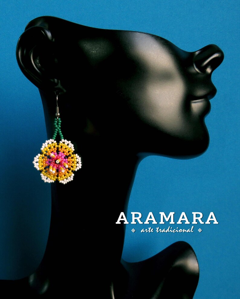 May include: A pair of beaded earrings with a pink, yellow, and white flower design. The earrings are hanging from a black mannequin's ear. The text "ARAMARA arte tradicional" is visible below the mannequin.