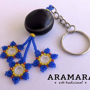 K&ouml;nnte beinhalten: Ein Schl&uuml;sselanh&auml;nger aus einer schwarzen Samenkugel mit drei blauen, gelben und wei&szlig;en Perlenblumen. Der Schl&uuml;sselanh&auml;nger ist mit "ARAMARA arte tradicional" beschriftet.
