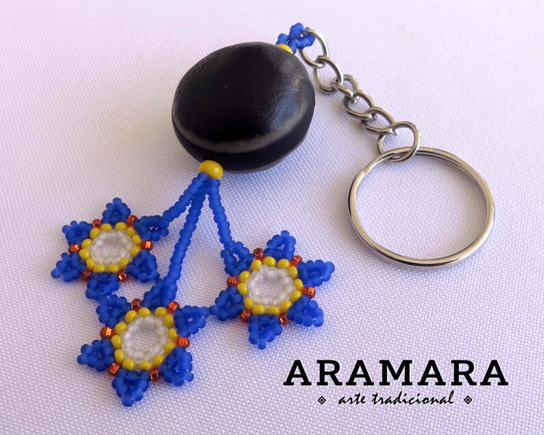K&ouml;nnte beinhalten: Ein Schl&uuml;sselanh&auml;nger aus einer schwarzen Samenkugel mit drei blauen, gelben und wei&szlig;en Perlenblumen. Der Schl&uuml;sselanh&auml;nger ist mit "ARAMARA arte tradicional" beschriftet.