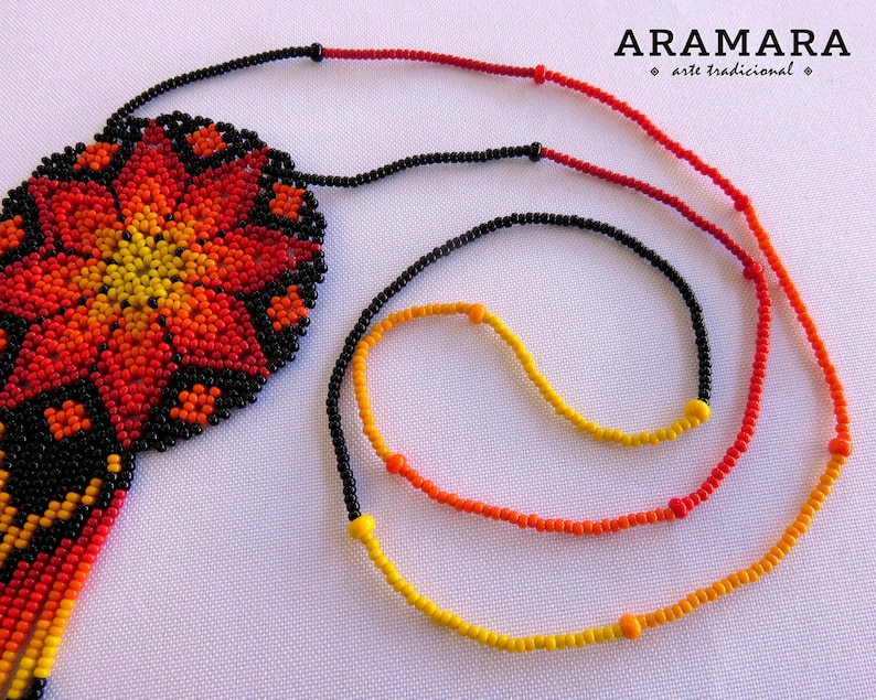 May include: A beaded necklace with a large, colorful flower pendant. The pendant is made of black, red, orange, and yellow beads. The necklace is made of black, red, orange, and yellow beads. The necklace is called "Aramara" and is described as "arte tradicional".
