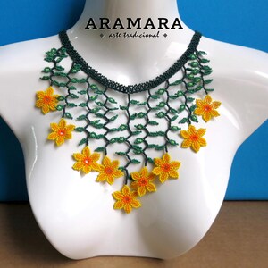 Op de afbeelding: Een kralenketting met een bloemenontwerp. De ketting heeft gele en oranje bloemkralen, groene wijnstokachtige kralen en een donkergroene kralen kraag. De ketting wordt getoond op een witte mannequin. De tekst "ARAMARA arte tradicional" staat bovenaan.
