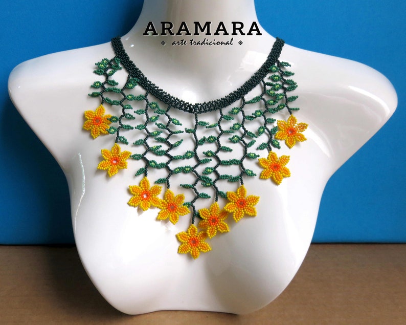 Op de afbeelding: Een kralenketting met een bloemenontwerp. De ketting heeft gele en oranje bloemkralen, groene wijnstokachtige kralen en een donkergroene kralen kraag. De ketting wordt getoond op een witte mannequin. De tekst "ARAMARA arte tradicional" staat bovenaan.