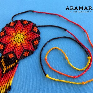 May include: A beaded necklace with a large, round pendant featuring a colorful geometric design in shades of red, orange, and yellow. The pendant is attached to a black beaded cord with a fringe of red, orange, and yellow beads.