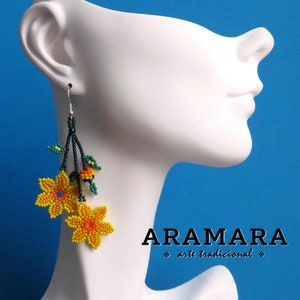 Op de afbeelding: Gebeelde bloemvormige oorbellen in geel en oranje, met groene bladeren en donkerblauwe stelen. De oorbellen worden getoond op een witte mannequinhoofd tegen een blauwe achtergrond. De tekst "ARAMARA arte tradicional" is zichtbaar.