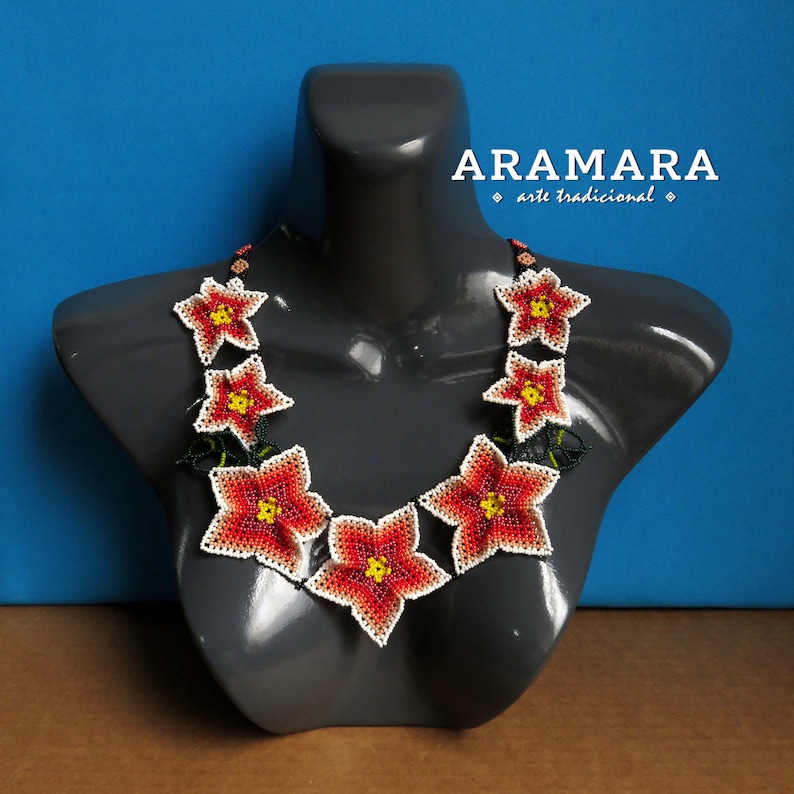 May include: A beaded necklace with red, yellow, and white flowers. The necklace is on a gray mannequin and the text "ARAMARA arte tradicional" is above the necklace.