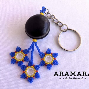 K&ouml;nnte beinhalten: Ein Schl&uuml;sselanh&auml;nger mit einer blauen und gelben Perlenblume, einem schwarzen Samenkapsel-Anh&auml;nger und einem silbernen Schl&uuml;sselring. Der Schl&uuml;sselanh&auml;nger ist mit "ARAMARA arte tradicional" beschriftet.