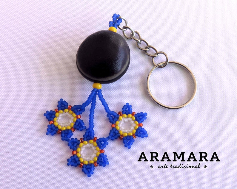 K&ouml;nnte beinhalten: Ein Schl&uuml;sselanh&auml;nger mit einer blauen und gelben Perlenblume, einem schwarzen Samenkapsel-Anh&auml;nger und einem silbernen Schl&uuml;sselring. Der Schl&uuml;sselanh&auml;nger ist mit "ARAMARA arte tradicional" beschriftet.
