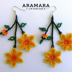 Op de afbeelding: Handgemaakte kralen oorbellen met gele en oranje bloemmotieven, groene bladeren en oranje accenten. De oorbellen hangen aan zilveren haken en hebben de tekst "ARAMARA arte tradicional".