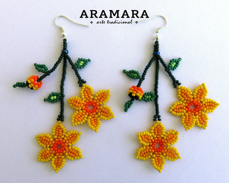 Op de afbeelding: Handgemaakte kralen oorbellen met gele en oranje bloemmotieven, groene bladeren en oranje accenten. De oorbellen hangen aan zilveren haken en hebben de tekst "ARAMARA arte tradicional".