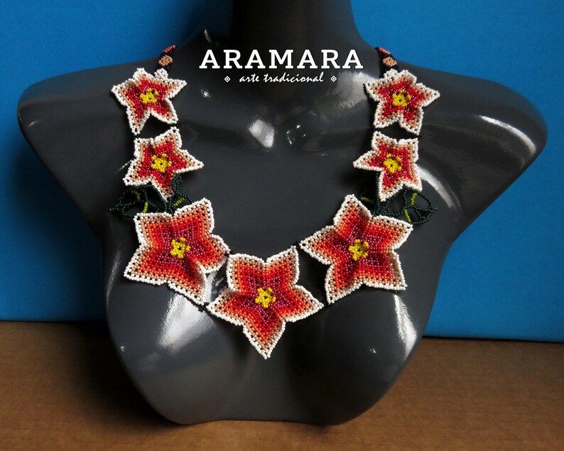 May include: A necklace made of red, white, and yellow beaded flowers. The necklace is on a gray mannequin and the background is blue. The text "ARAMARA arte tradicional" is on the necklace.