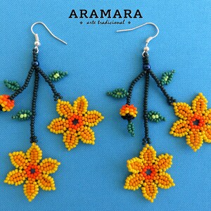 Op de afbeelding: Handgemaakte kralen oorbellen met gele en oranje bloemmotieven, groene bladeren en zwarte stelen. De oorbellen hangen aan zilveren haken. De tekst "ARAMARA arte tradicional" staat bovenaan.