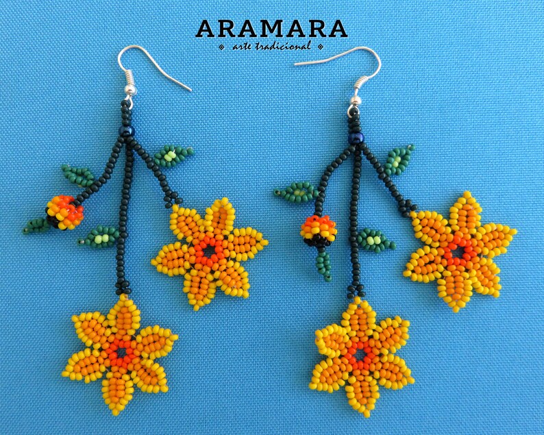 Op de afbeelding: Handgemaakte kralen oorbellen met gele en oranje bloemmotieven, groene bladeren en zwarte stelen. De oorbellen hangen aan zilveren haken. De tekst "ARAMARA arte tradicional" staat bovenaan.