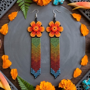 Könnte beinhalten: Ein Paar Perlenohrringe mit einem Blumenmuster in Orange und Schwarz. Die Ohrringe haben eine lange, gefranste, regenbogenfarbene Quaste. Die Ohrringe werden auf einer grauen Oberfläche mit orangefarbenen Blütenblättern und kleinen Schalen mit Perlen präsentiert.