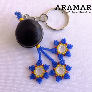 K&ouml;nnte beinhalten: Ein Schl&uuml;sselanh&auml;nger aus schwarzer Saatperle mit drei blauen, gelben und wei&szlig;en Perlenblumenanh&auml;ngern. Der Schl&uuml;sselanh&auml;nger ist mit "ARAMARA arte tradicional" beschriftet.