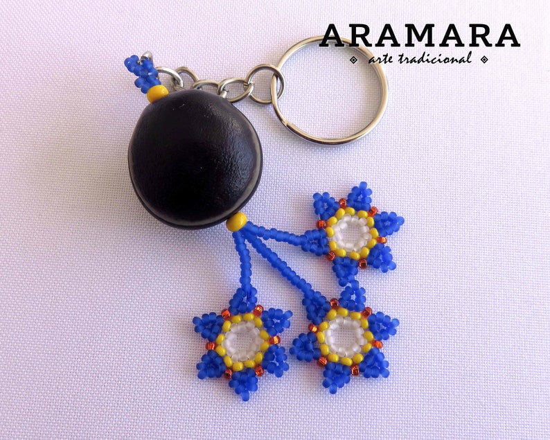 K&ouml;nnte beinhalten: Ein Schl&uuml;sselanh&auml;nger aus schwarzer Saatperle mit drei blauen, gelben und wei&szlig;en Perlenblumenanh&auml;ngern. Der Schl&uuml;sselanh&auml;nger ist mit "ARAMARA arte tradicional" beschriftet.