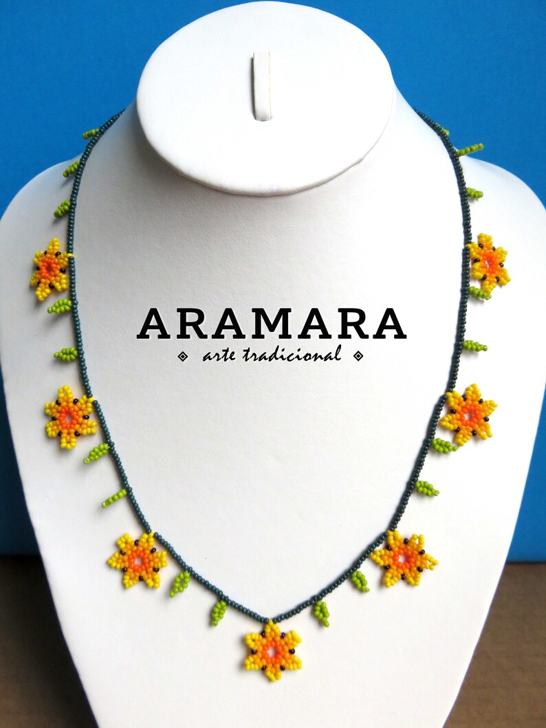 May include: A beaded necklace with a floral design. The necklace is made with dark blue beads and features yellow and orange flowers with green leaves. The necklace has the text "ARAMARA arte tradicional" on a white background.