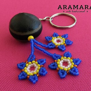 K&ouml;nnte beinhalten: Eine braune und schwarze Samenkapsel mit einem silbernen Schl&uuml;sselring. Drei blaue Perlenblumen mit gelben und wei&szlig;en Zentren h&auml;ngen an der Samenkapsel. Die Blumen befinden sich auf einem rosa Hintergrund.  "ARAMARA arte tradicional" ist in wei&szlig; auf schwarzem Hintergrund geschrieben.