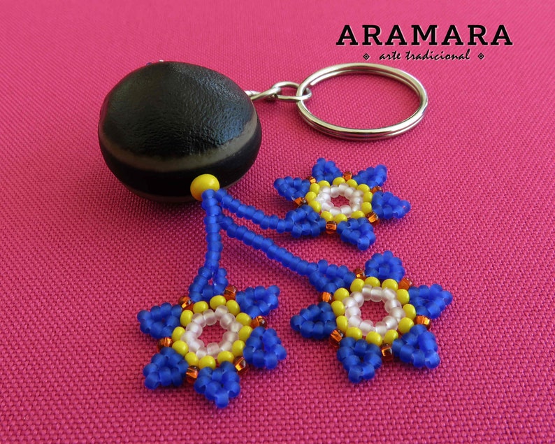K&ouml;nnte beinhalten: Eine braune und schwarze Samenkapsel mit einem silbernen Schl&uuml;sselring. Drei blaue Perlenblumen mit gelben und wei&szlig;en Zentren h&auml;ngen an der Samenkapsel. Die Blumen befinden sich auf einem rosa Hintergrund.  "ARAMARA arte tradicional" ist in wei&szlig; auf schwarzem Hintergrund geschrieben.